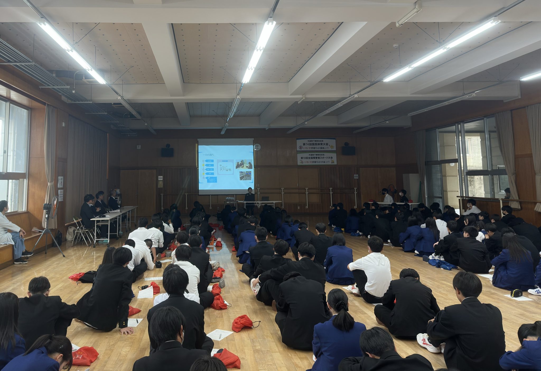 水戸商業高等学校の情報ビジネス課の約160名の生徒の前で、両行出身者の弊社社員が講演を行っている様子です。 srcset=